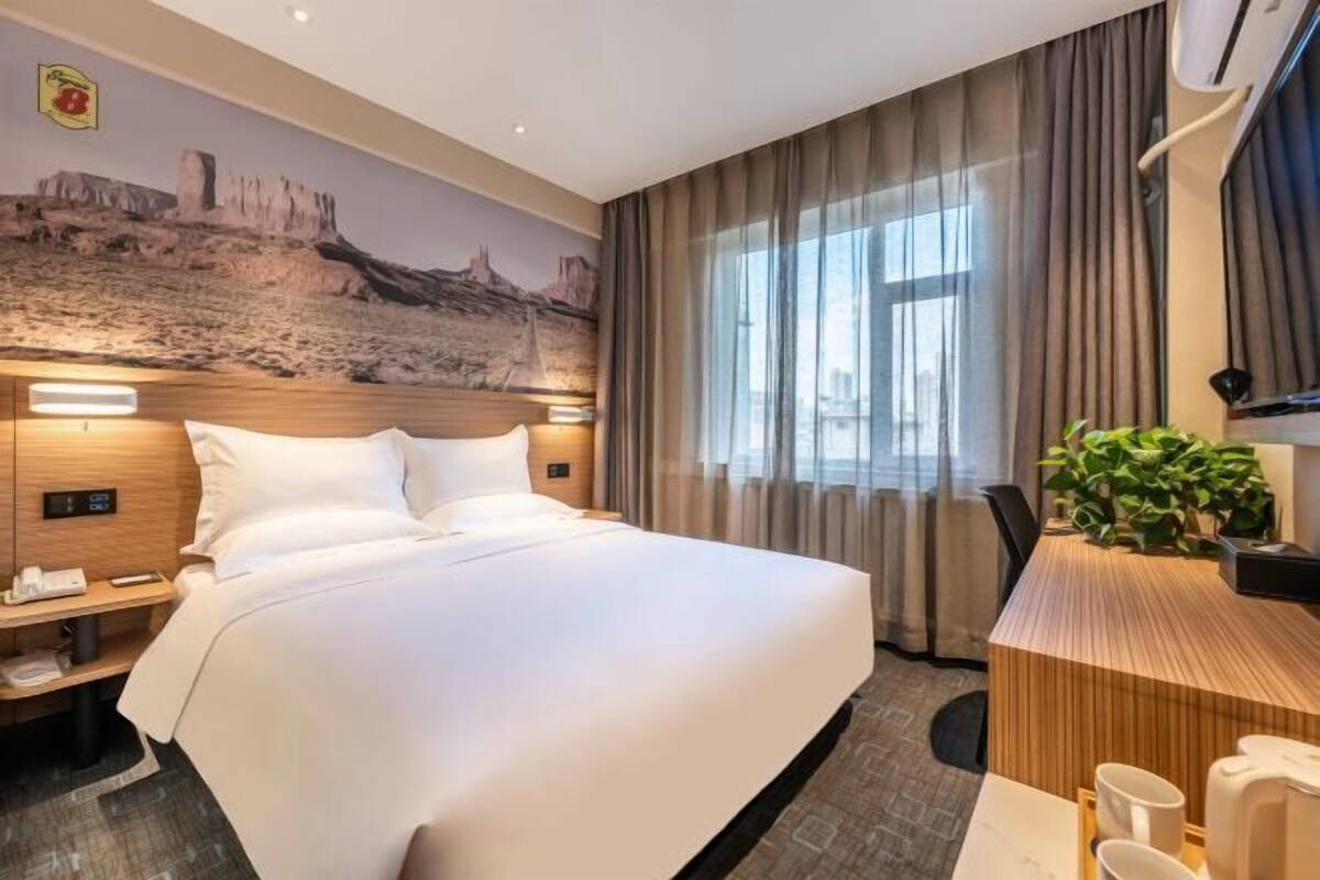 A hotel room with a large bed, a desk with chairs, a plant, and a cityscape mural on the wall.