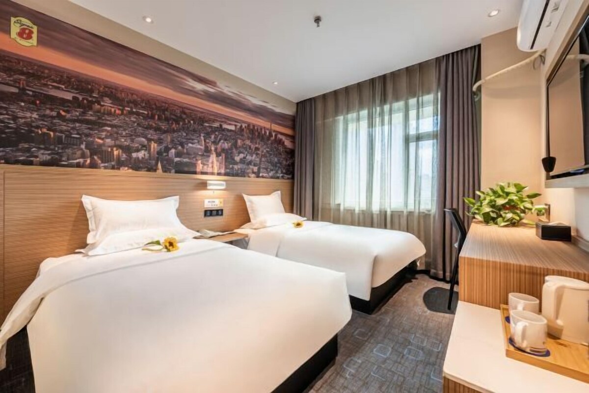 A hotel room with two beds, a desk, a chair, a TV, and a large mural on the wall.