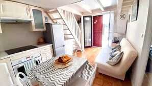 Une cuisine compacte avec des meubles blancs, un réfrigérateur et un coin repas avec une table pour deux personnes. Un escalier mène à un espace de vie comprenant un canapé et une lampe.