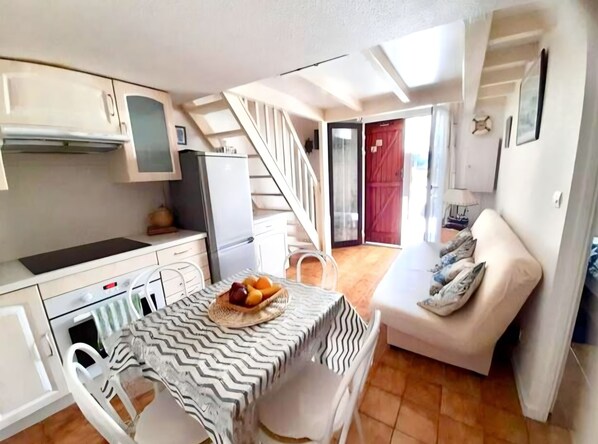 Una cocina compacta con armarios blancos, un refrigerador y un comedor con una mesa dispuesta para dos personas. Una escalera conduce a una sala de estar con un sofá y una lámpara.