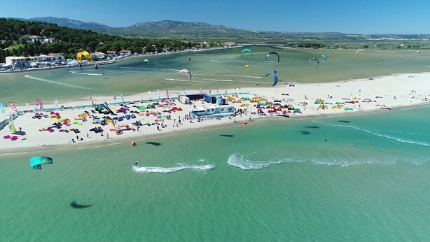 Vue aérienne d’une plage où l’on aperçoit de nombreuses voiles de parachute multicolores, une plage de sable et une mer d’un bleu limpide.