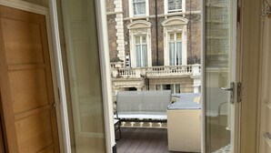 Ein Schlafzimmer mit großem Fenster, einem Bett mit weißer Bettwäsche und Blick auf ein Gebäude mit einem Balkon.