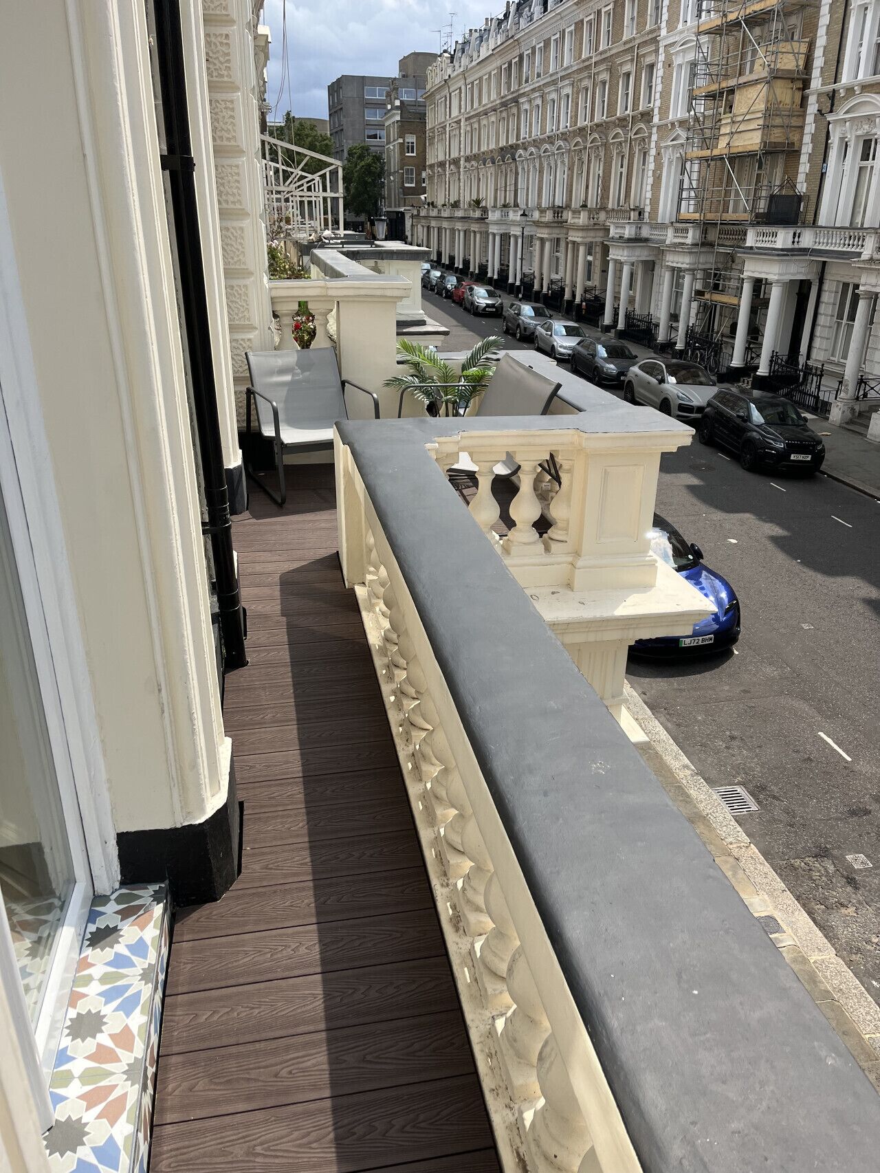 A balcony with a wooden floor, white balustrade, and a view of parked cars and buildings.