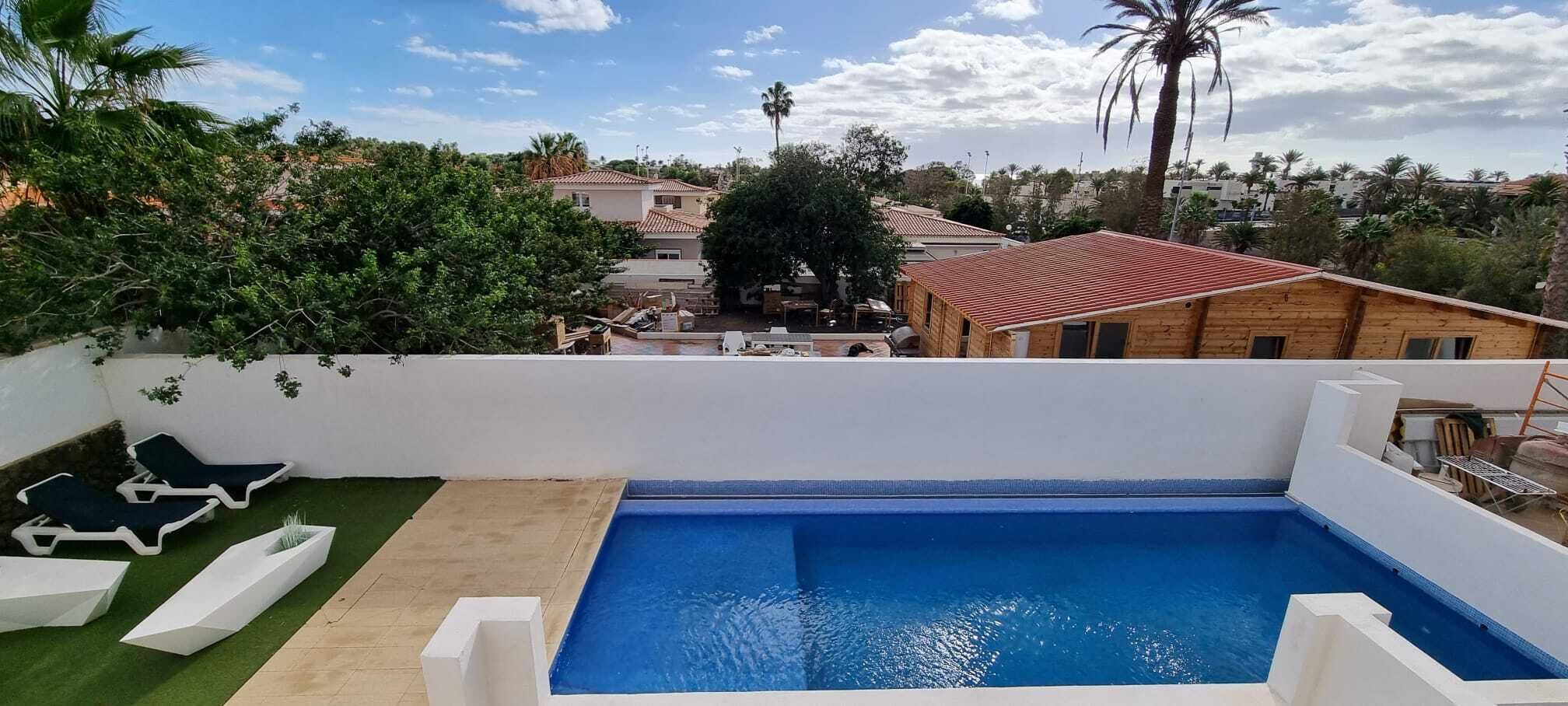 Una piscina en la azotea con sillones blancos, un pequeño jardín y vistas de edificios y árboles circundantes.