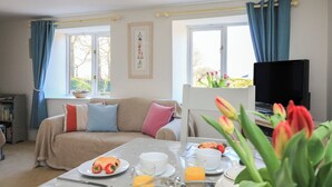 A cozy room with a sofa, a dining table with breakfast, a TV, and windows with curtains. - Tinkers Cottage (Kingsbridge)