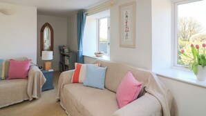A cozy living room with a beige sofa, a blue armchair, and a window with a view of greenery. - Tinkers Cottage (Kingsbridge)