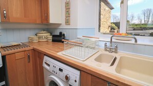 A kitchen with wooden cabinets, a Bosch washing machine, a sink, and a window with a view of a stone building and trees. - Tinkers Cottage (Kingsbridge)