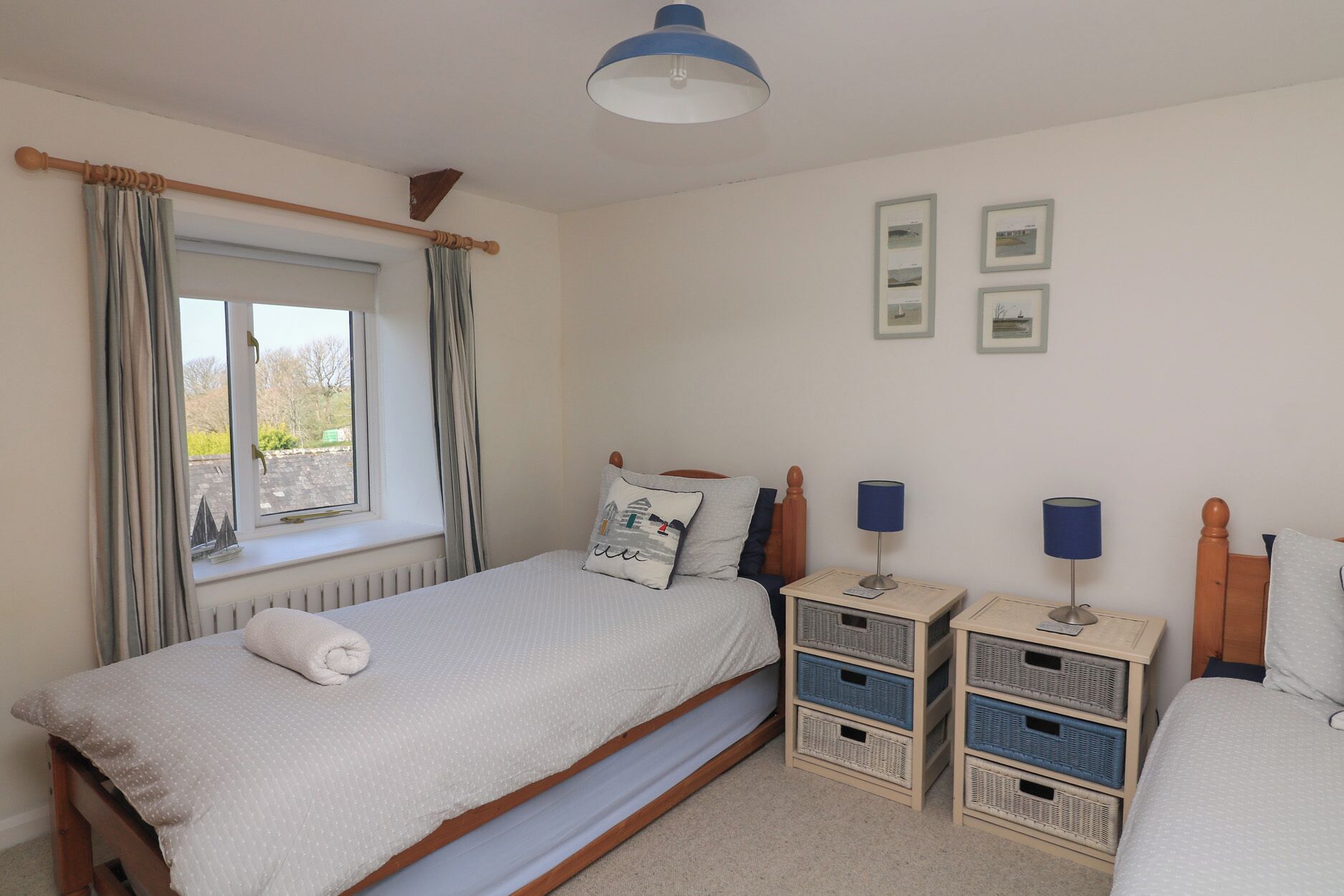Un dormitorio con cama, ventana con cortinas, mesita de noche con lámparas y cuadros en la pared.