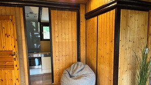 Una habitación con paredes de madera, un área de cocina visible a través de una puerta abierta y una zona de estar con un sillón de mimbre y un reposapiés.