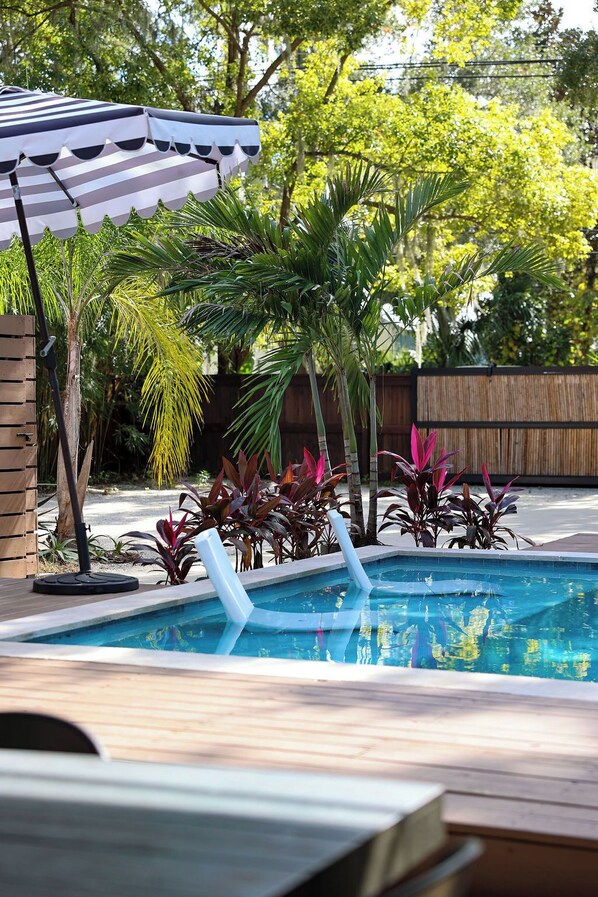 A swimming pool with a wooden deck, surrounded by tropical plants and a striped umbrella.