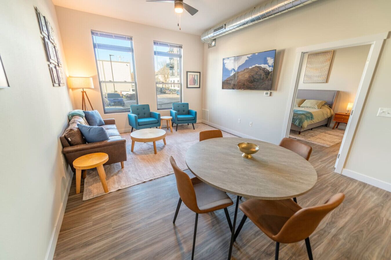 A modern living room with a sofa, two armchairs, a coffee table, and a dining table with chairs. There are large windows providing natural light and a view of the outside.