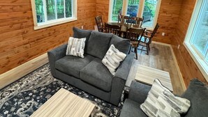 A cozy cabin interior with a gray sofa, a dining table with chairs, and large windows offering a view of the forest.
