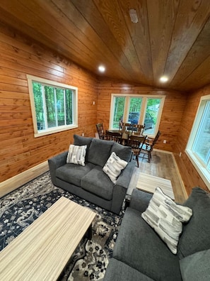 A cozy cabin interior with a gray sofa, a dining table with chairs, and large windows offering a view of the forest. - River Retreat Tiny Homes:Wood Sauna/Rec Room/River (Willard)