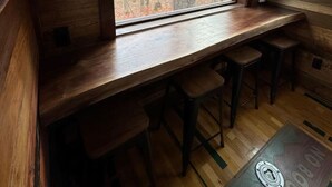 A rustic cabin interior with a wooden bar area, a large window offering a view of a forest, and a wooden floor.