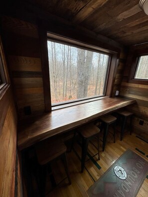 A rustic cabin interior with a wooden bar area, a large window offering a view of a forest, and a wooden floor. - River Retreat Tiny Homes:Wood Sauna/Rec Room/River (Willard)