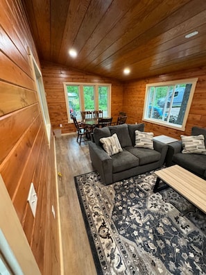 A cozy cabin-style room with a wooden interior, a gray sofa set, a patterned rug, and a dining area with a table and chairs.