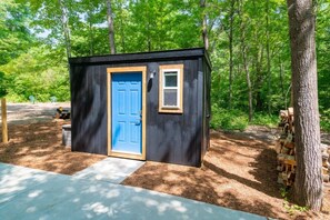 A small, dark wooden shed with a blue door and a small window, surrounded by trees and a concrete path.