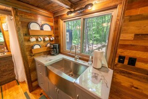 Interior de una cabaña de madera con cocina, que incluye un lavamanos, una cafetera y un ventanal con vistas al bosque.