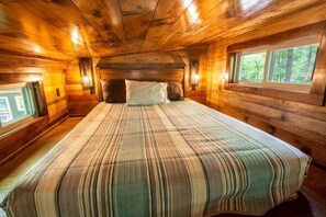 A cabin-style room with a large bed, wooden walls, and windows offering a view of the forest.