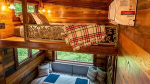 A cabin interior with a loft bed, a bench seat, and a window overlooking greenery.