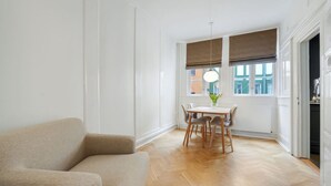 A room with a beige sofa, a wooden dining table with chairs, a hanging light fixture, and a window with blinds. - Beautiful apartment in the center of KBH (København)