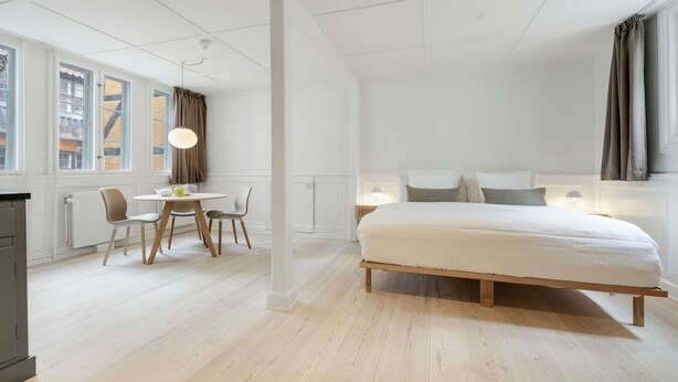 A modern bedroom with a large bed, a dining area with a table and chairs, and a view of the city through large windows. - Small apartment in the heart of KBH (København)