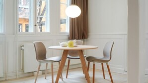 A dining area with a round table, four chairs, a pendant light, and a view of buildings through large windows. - Small apartment in the heart of KBH (København)