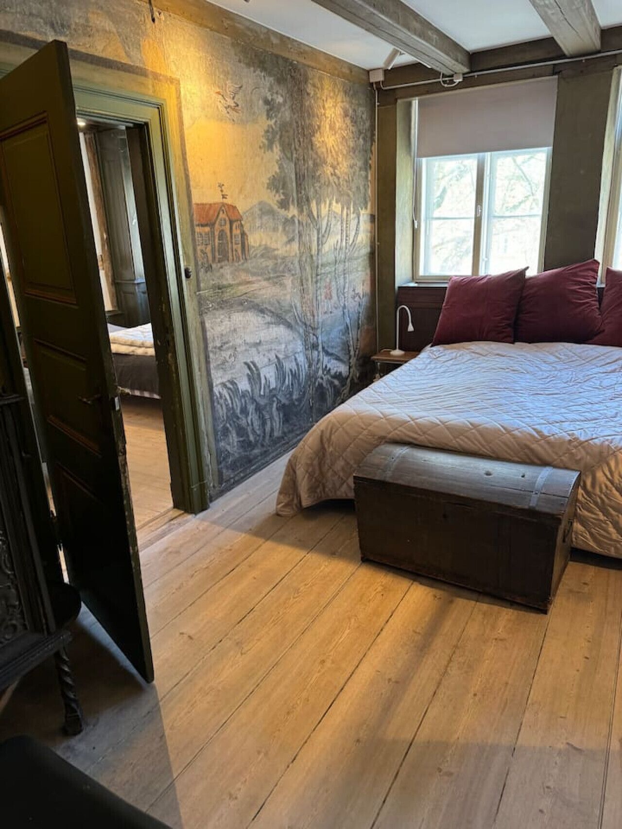 A bedroom with a bed, a bench, a mural, and a window.