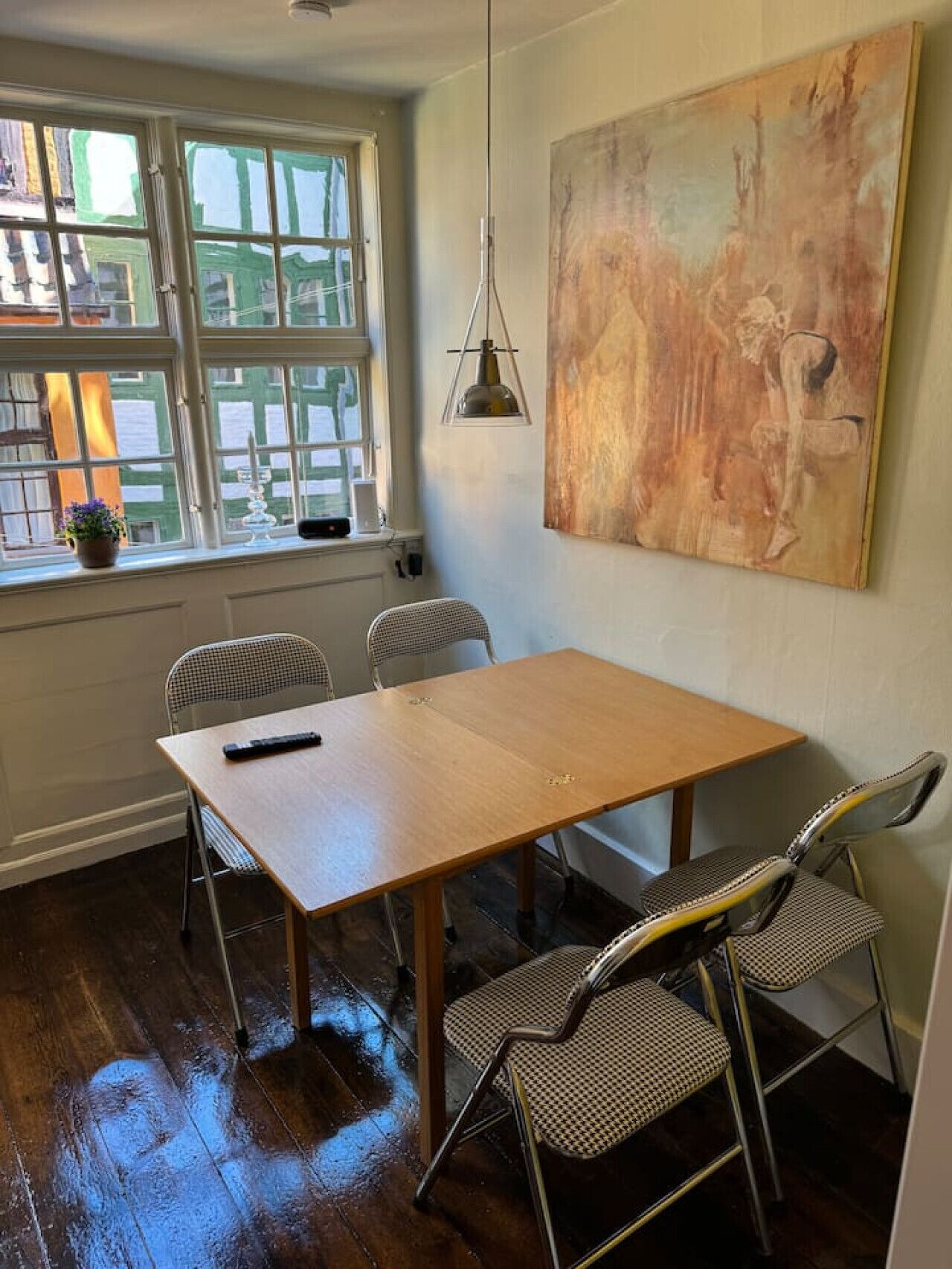 A room with a wooden table, four chairs, a painting on the wall, a window with a view, and a pendant light.