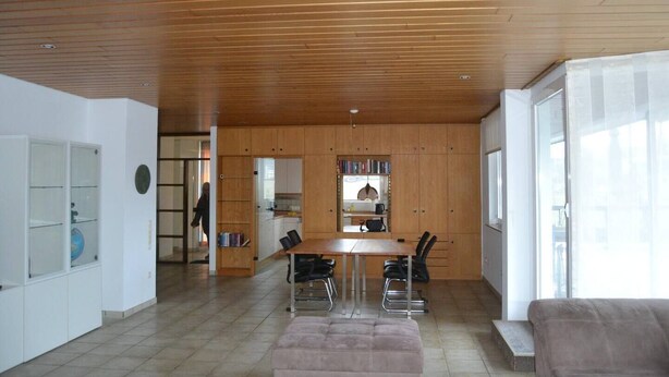 A modern living space with a wooden ceiling, a dining area with a table and chairs, a sofa, and a kitchen area visible through a glass door. - PentArtHouse Apartment (Burscheid)