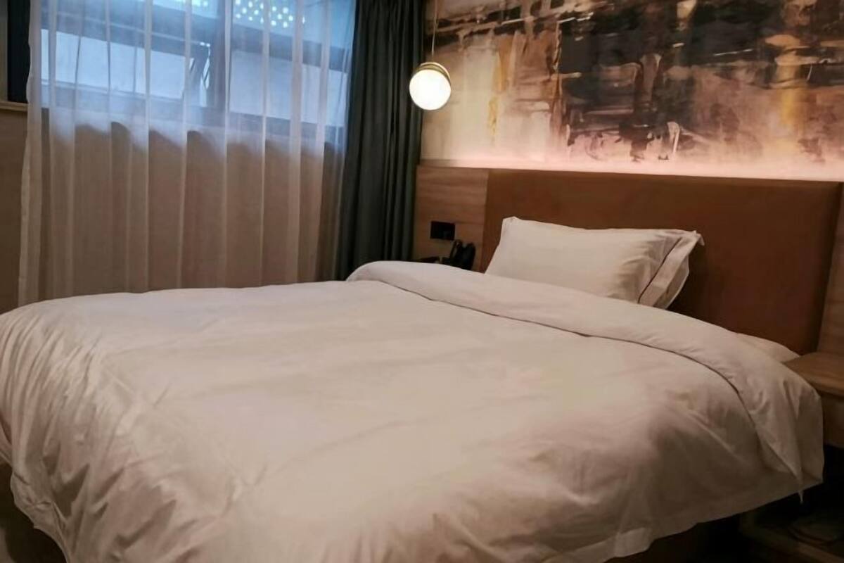 A hotel room with a large bed, a headboard with a light fixture, and a view through sheer curtains.