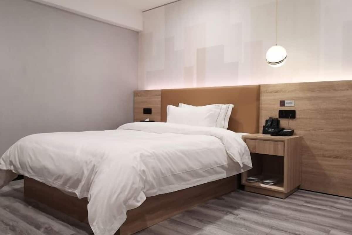 A modern hotel room with a wooden headboard, a white bedspread, a bedside table with a lamp, and a built-in shelf.