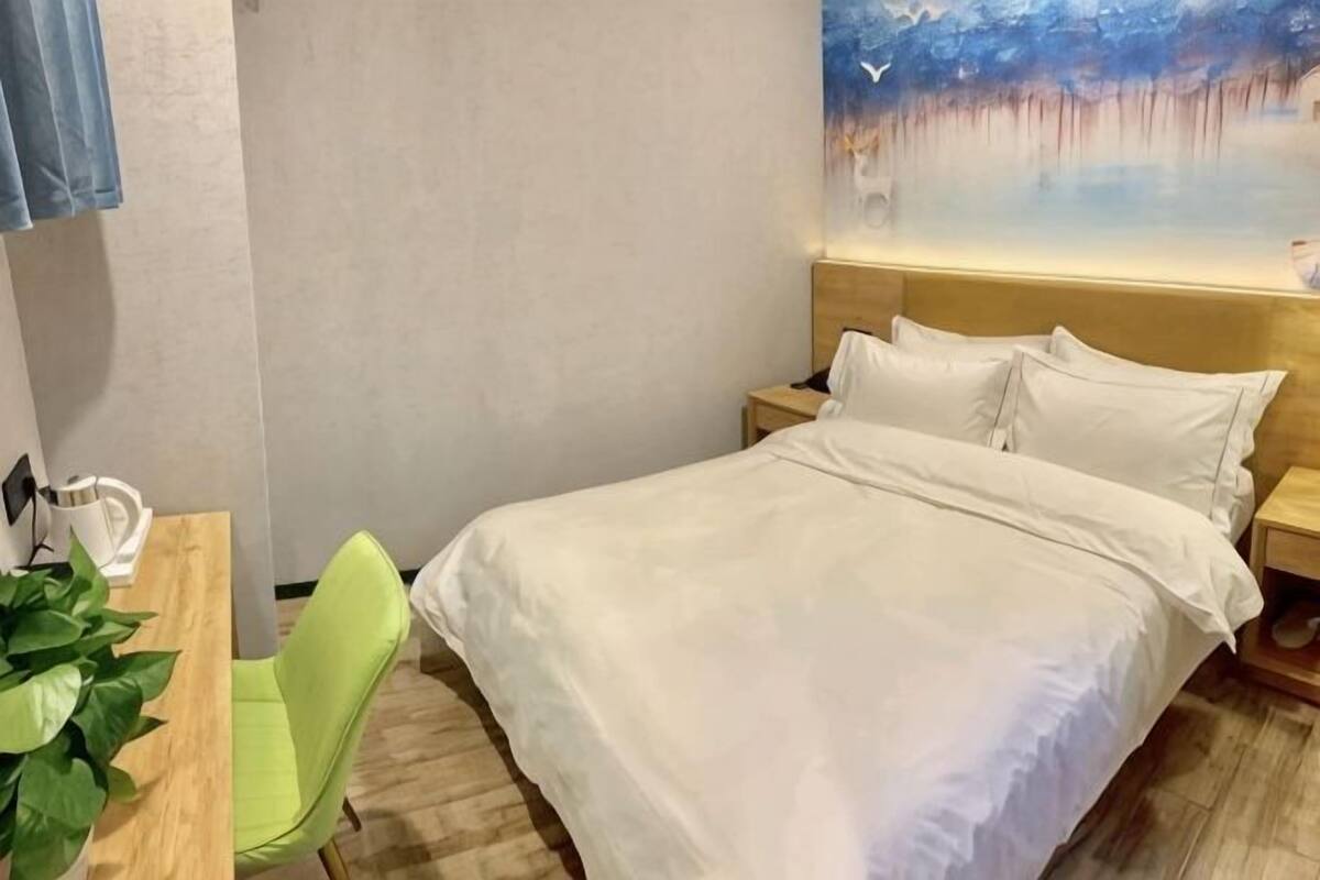 A hotel room with a bed, a desk with a chair, and a plant.