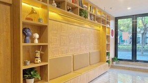 A modern hotel reception area with wooden shelving, potted plants, and a large window offering a view of the outdoors.