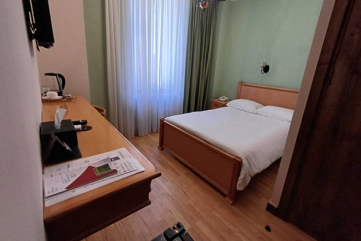 Une chambre d’hôtel avec un bureau en bois, un lit, une table de chevet, un téléphone et une fenêtre avec des rideaux.