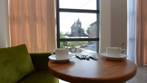 A room with a green sofa, a wooden table with two white cups and saucers, and a window with a view of a church. - Old Avenue Hotel (Gyumri)