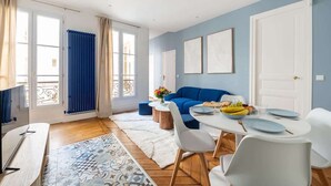 Ein modernes Wohnzimmer mit einem blauen Sofa, einem Esstisch für vier Personen und einem großen Fenster mit Vorhängen.