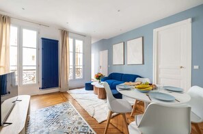 Ein modernes Wohnzimmer mit einem blauen Sofa, einem Esstisch für vier Personen und einem großen Fenster mit Vorhängen.