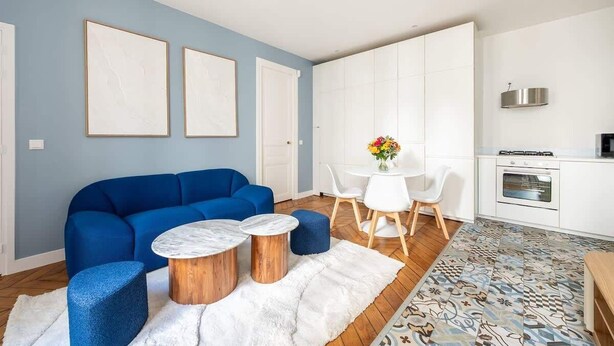 Ein modernes Wohnzimmer mit einem blauen Sofa, runden Couchtischen, einem weißen Teppich und einem Essbereich mit weißem Tisch und Stühlen.