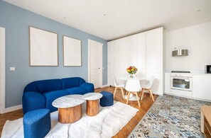 Ein modernes Wohnzimmer mit einem blauen Sofa, runden Couchtischen, einem weißen Teppich und einem Essbereich mit weißem Tisch und Stühlen.