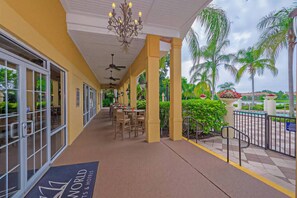 A covered walkway with tables and chairs, a ceiling fan, and a view of palm trees and a pool.