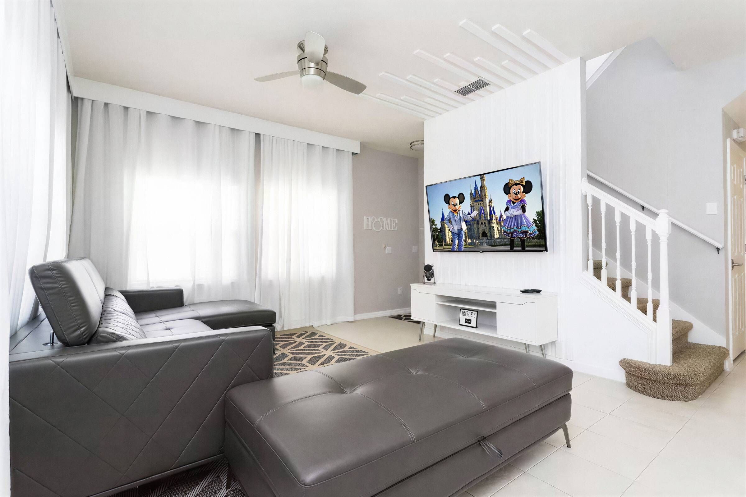 A modern living room with a grey leather sofa, a flat-screen TV mounted on a white wall, and a staircase leading to an upper level.