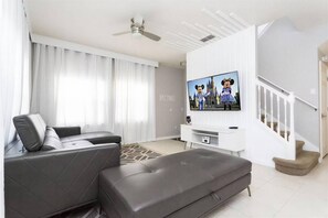 A modern living room with a grey leather sofa, a flat-screen TV mounted on a white wall, and a staircase leading to an upper level. - 4BR Pool Home Near Disney at Encantada (Kissimmee)