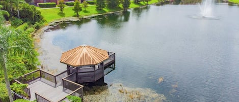 Vista de un lago con muelle de madera y un pabellón, rodeado de vegetación y edificios residenciales.