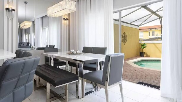 Un comedor moderno con una mesa blanca y sillas grises, una araña y vistas a una piscina.