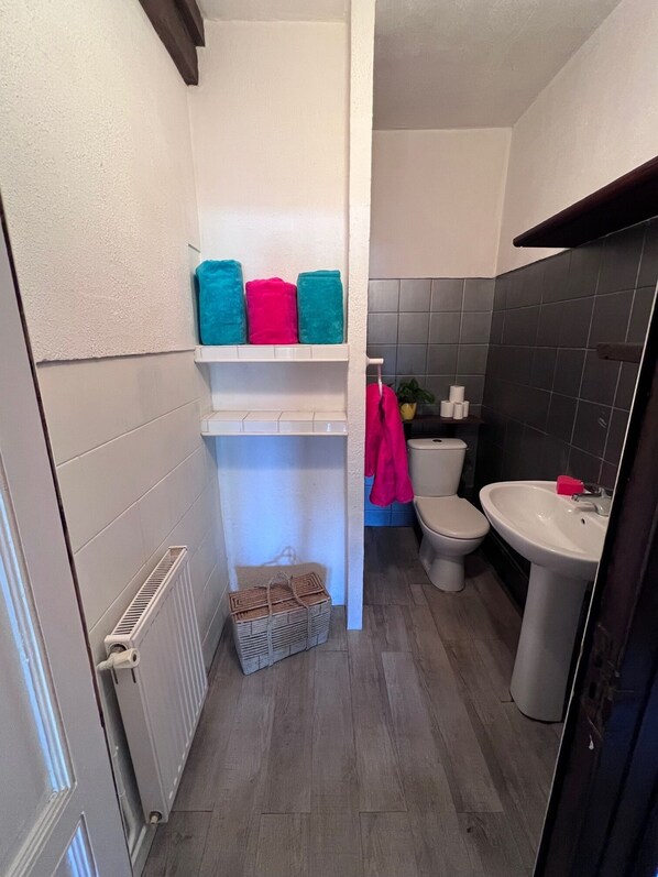 A bathroom with a white radiator, a white sink, and a toilet. There are towels on a shelf and a basket on the floor.