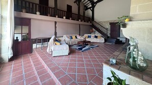 A spacious living area with a high ceiling, terracotta tiled floor, a sofa set, a staircase, and a view of the upper floor.