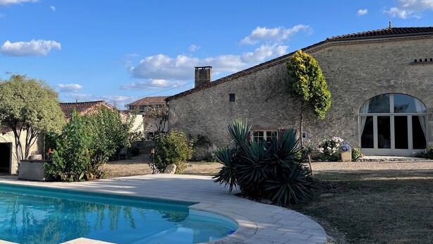A stone building with a swimming pool, surrounded by greenery and a clear sky.