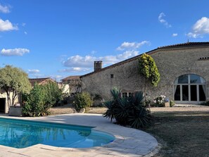 A stone building with a swimming pool, surrounded by greenery and a clear sky.