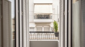 Un balcón estrecho con una maceta, contraventanas blancas y vistas a un edificio con varias ventanas y balcones.